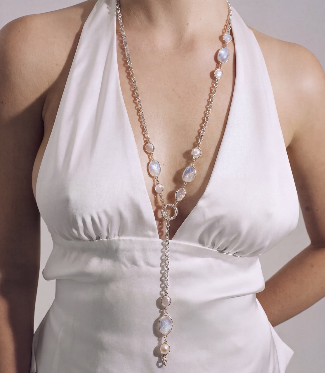 A close-up of a model wearing a long double-strand lariat necklace with baroque pearls and silver-toned chain links, styled with a white halter-neck dress.
