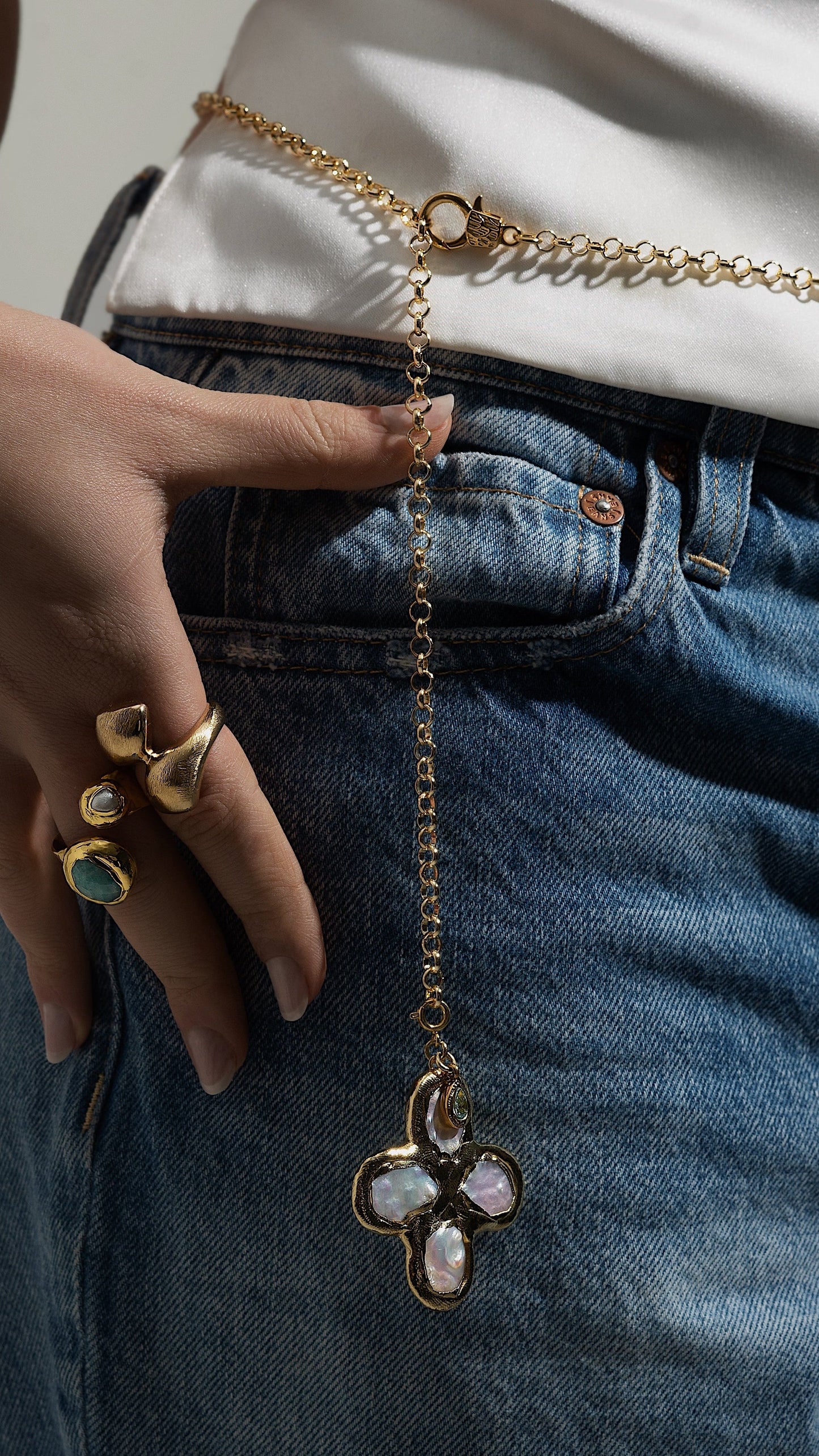 Person wearing a gold chain belt with a floral pendant on blue jeans against a neutral background