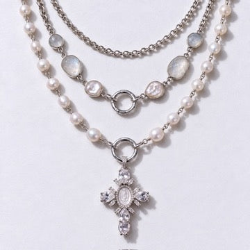 A layered silver-tone necklace featuring white pearl beads, iridescent oval stones, and a dangling cross pendant embellished with clear rhinestones, displayed on a light gray background.