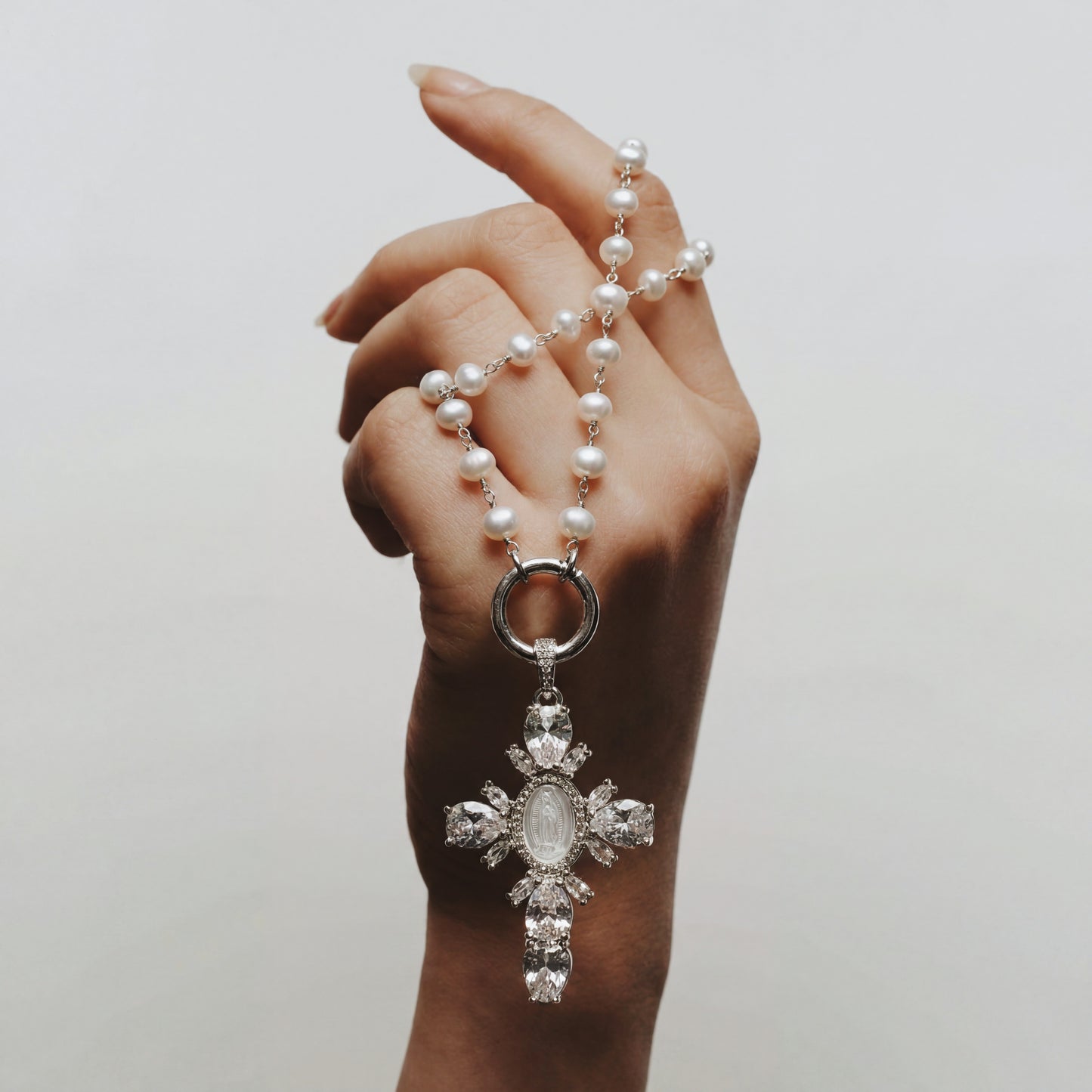 Hand holding a pearl necklace with a decorative cross pendant against a plain background