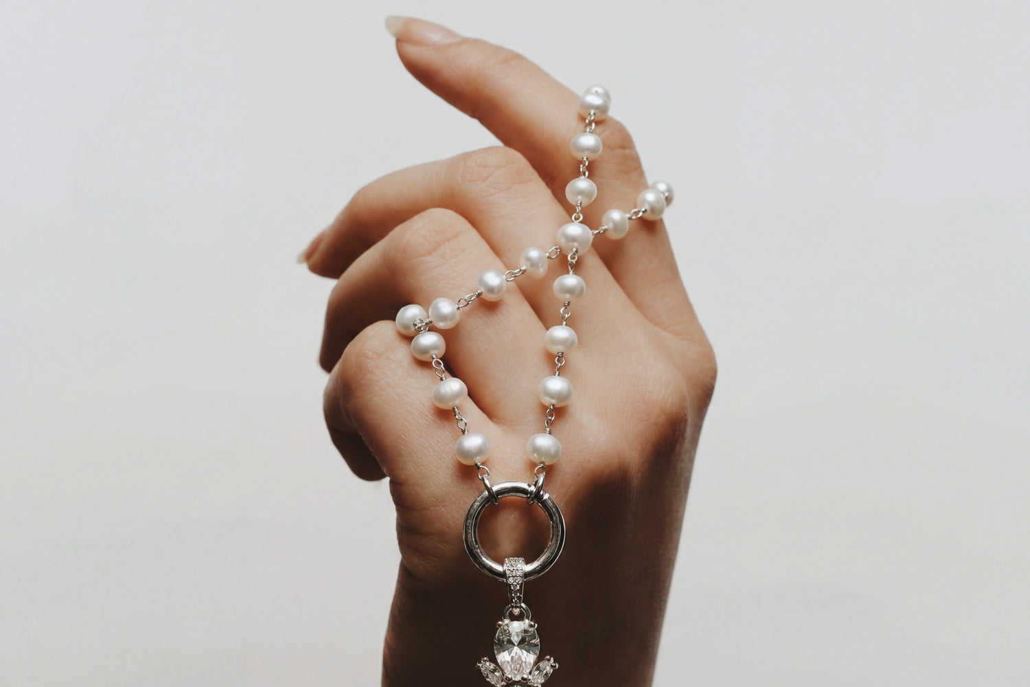Hand holding a pearl necklace with a decorative cross pendant against a plain background