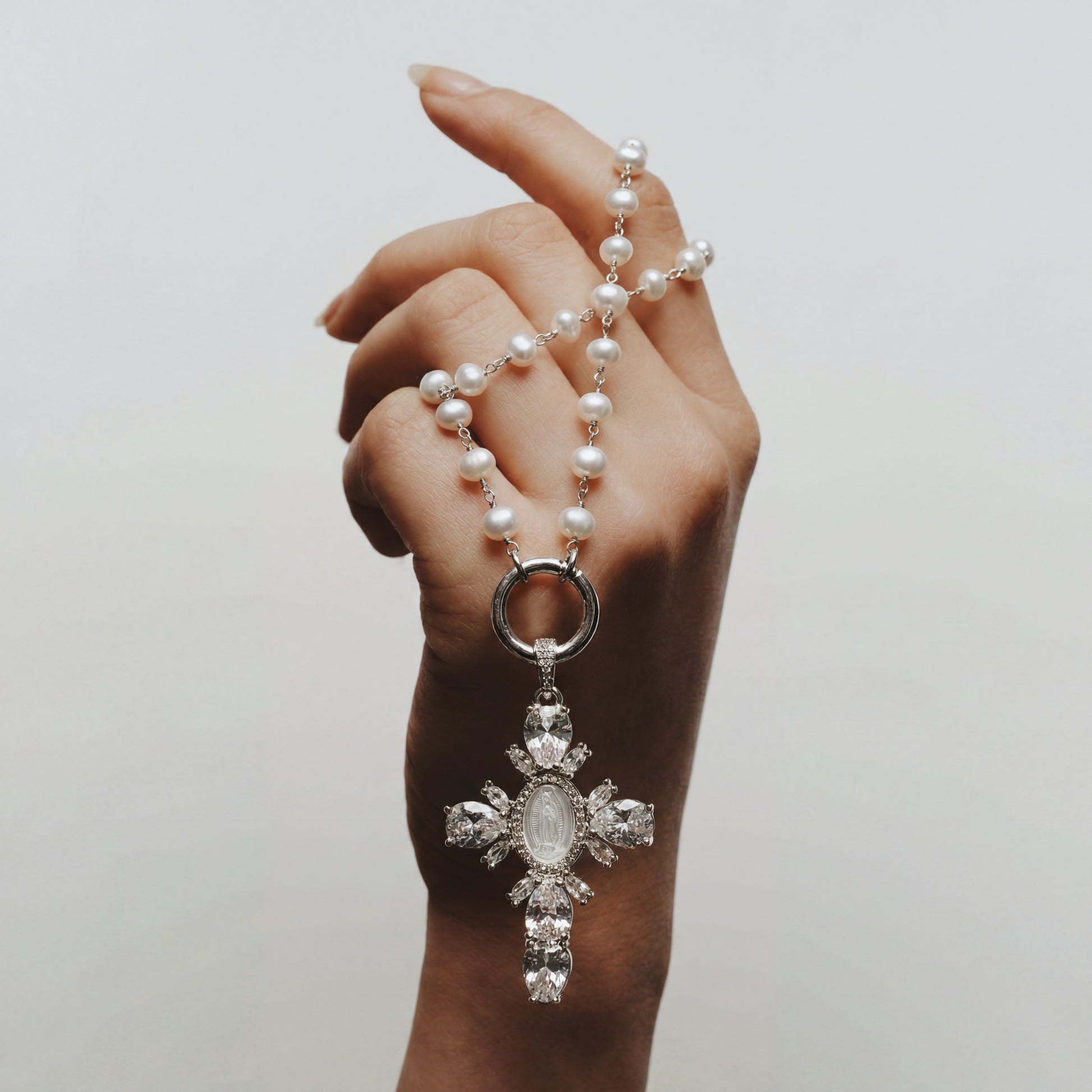 Hand holding a pearl necklace with a decorative cross pendant against a plain background