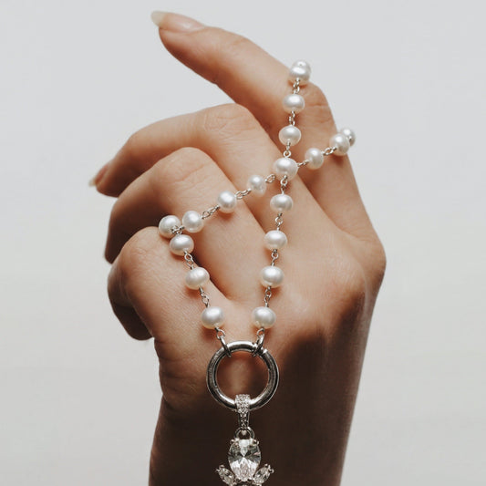 Hand holding a pearl necklace with a decorative cross pendant against a plain background