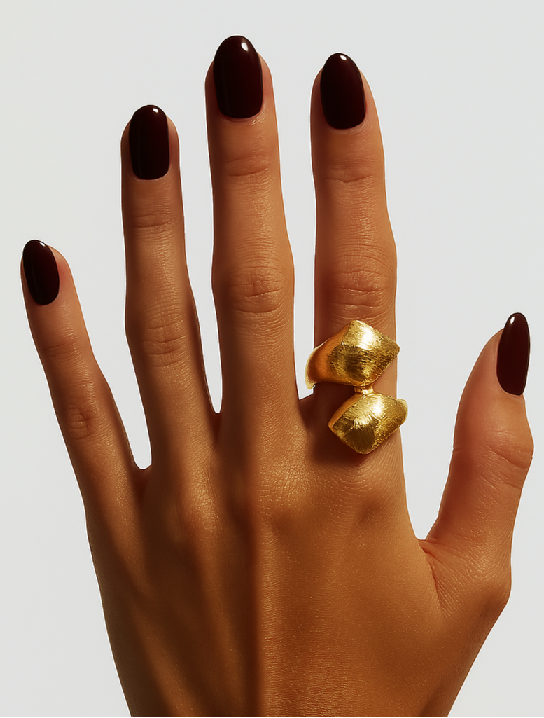 Hand wearing a gold ring with dark nail polish on a light background