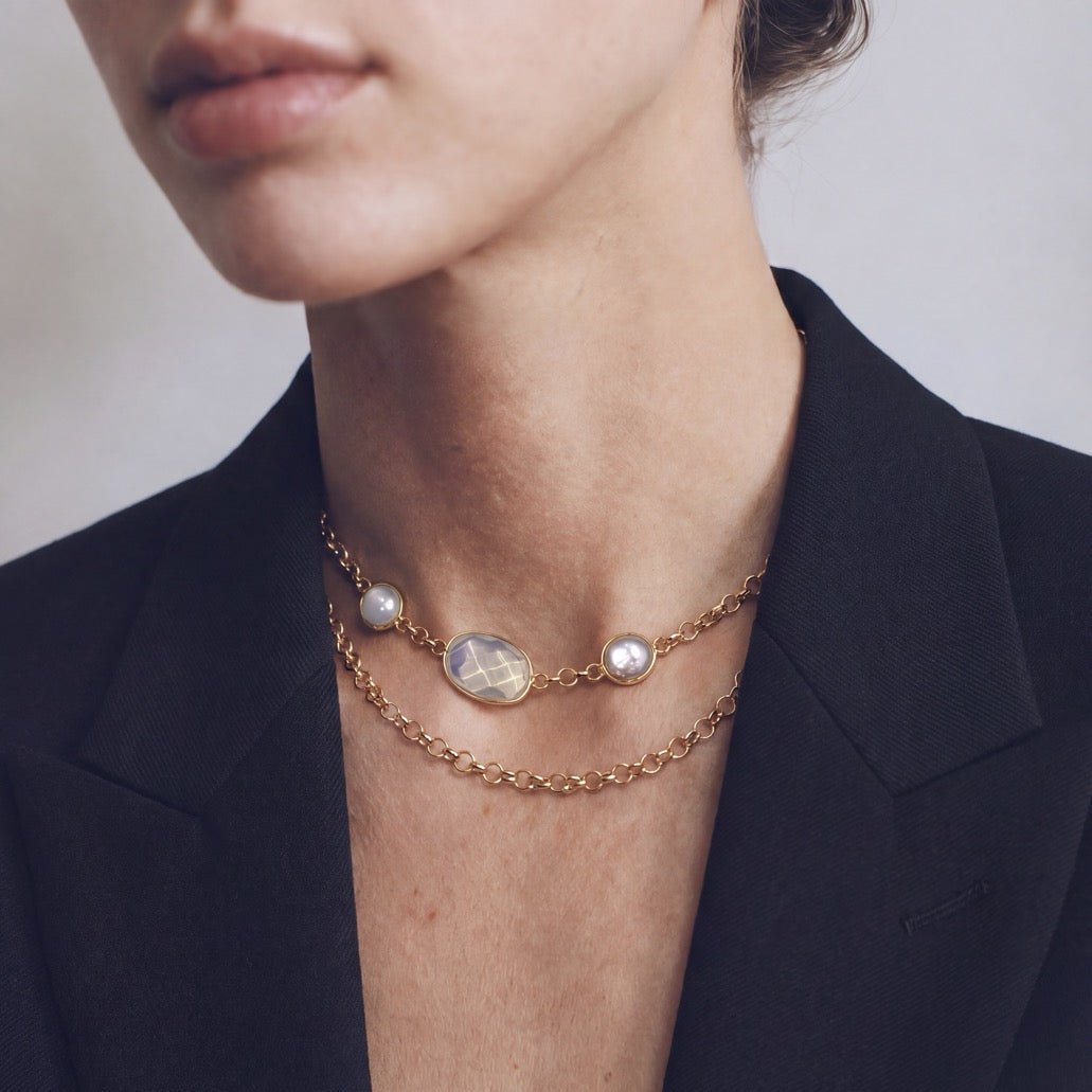 A close-up of a model wearing a double-layered gold chain necklace with white pearls and a central iridescent moonstone pendant, styled with a black blazer.
