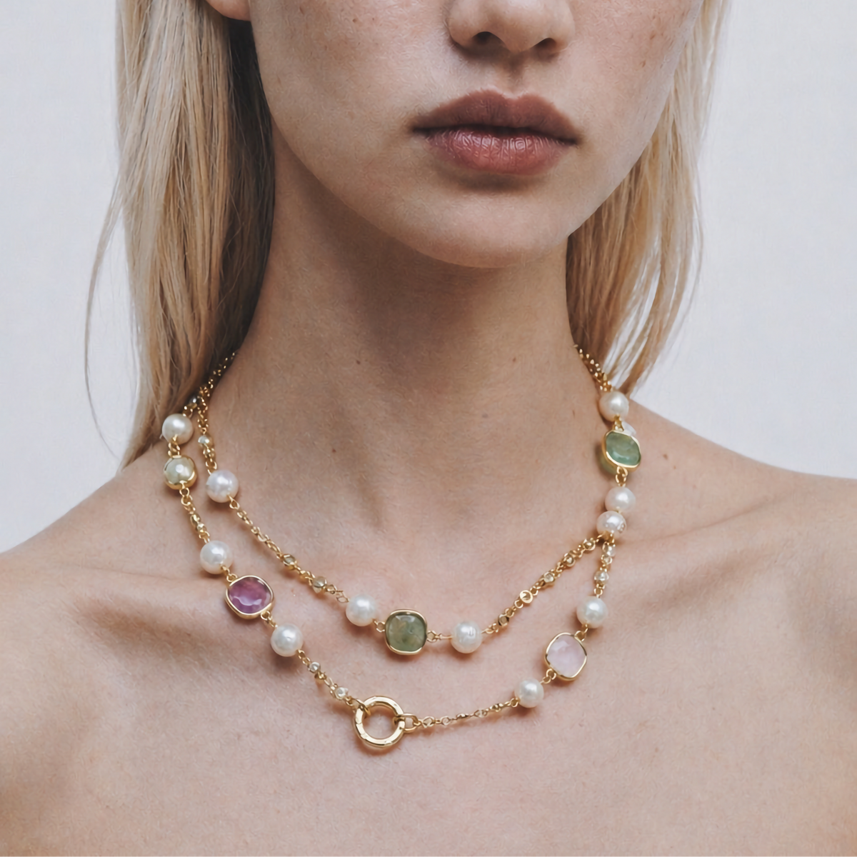 A close-up of a model wearing a delicate gold double-strand necklace with white pearls and small oval gemstone accents in green, pink, and purple, set against a plain light gray background.